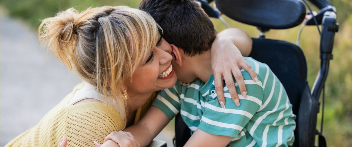 A smiling mother hugs her son, who sits in a wheelchair outdoors.