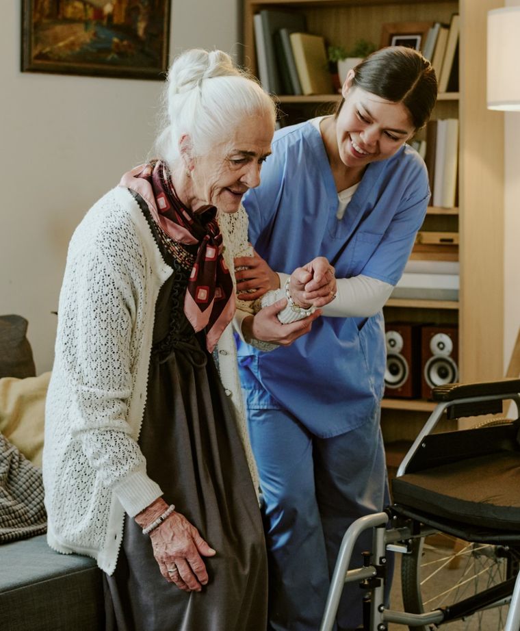 A home care aide smiles as she helps an older woman stand up from the sofa beside a wheelchair.