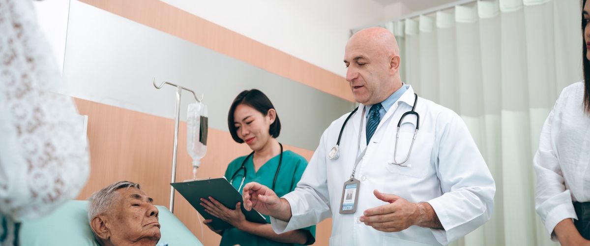 A doctor and nurse talk with an older man in a hospital bed while his family listens.