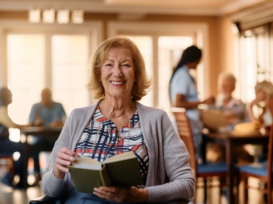 A smiling older woman in a wheelchair holds an open book in a sunny common room.